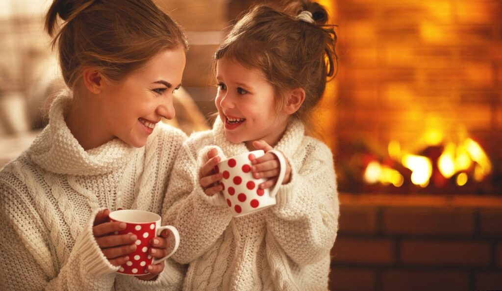 A mother and daughter wearing cozy sweaters, smiling and sharing a warm moment by the fireplace while holding mugs, celebrating Valentine's Day together